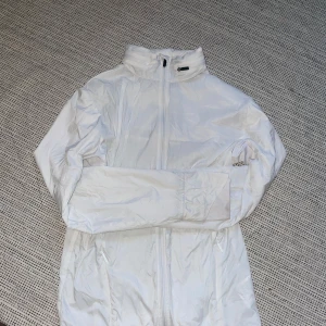 Vit vindjacka med dragkedja - Säljer en vit vindjacka i syntetmaterial. Jackan har en hög krage och är utrustad med en dragkedja framtill. Perfekt för vår och sommar när det blåser lite extra. Den har långa ärmar och en praktisk ficka med dragkedja på ryggen. Limited edition 