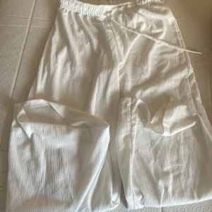 Vita byxor från Gina Tricot - Säljer ett par vita byxor från Gina Tricot i storlek S. De är gjorda i ett lätt och luftigt material, perfekt för varma dagar. Byxorna har en bekväm passform med resår i midjan och snörning för justering. Perfekta för en avslappnad och stilren look!