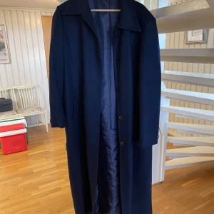 Mörkblå lång kappa -  Säljer en elegant mörkblå kappa i ull. Den har en klassisk design med knappar framtill och ett bälte i midjan för en snygg passform. Perfekt för höst och vinter, och den är långärmad för extra värme. Kappan är i bra skick och redo för nya äventyr! 