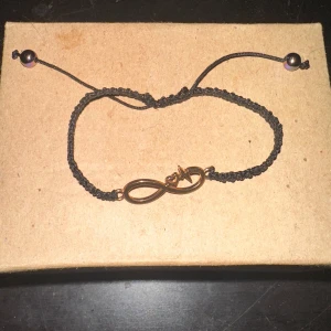 Svart armband med infinity-symbol - Säljer ett snyggt svart armband med en guldig infinity-symbol i mitten. Armbandet är justerbart med två små pärlor i ändarna. Perfekt för att ge en stilren touch till din outfit!