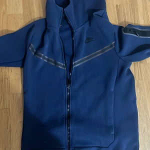 Blå hoodie från Nike - Säljer en snygg blå hoodie från Nike. Den har en dragkedja framtill och en cool design med svarta detaljer på ärmarna. Perfekt för både träning och vardag. Den är i bra skick och redo för en ny ägare!