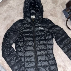 Svart dunjacka från Emporio Armani - Säljer en snygg svart dunjacka från Emporio Armani. Den har en stilren design med snygga svarta detaljer. Jackan är perfekt för hösten nu! Den är i bra skick förutom det lilla igensydda  hålet på baksidan som knappt syns!💓