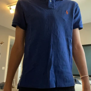 Blå pikétröja från Ralph Lauren - Säljer en snygg blå pikétröja från Ralph Lauren i storlek S. Den har en klassisk krage med knappar och det ikoniska broderade logotypen i orange på bröstet. Tröjan är i slim fit och passar perfekt för både vardag och fest. Materialet är mjukt och bekvämt, perfekt för sommardagar!