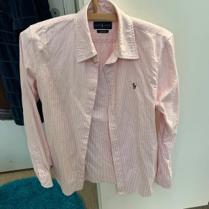 Rosa och vit randig skjorta från Ralph Lauren - Säljer en snygg och stilren rosa och vit randig skjorta från Ralph Lauren. Skjortan är i custom fit och har långa ärmar. Den är gjord i bomull och har klassiska knappar framtill. Perfekt för både vardag och fest!