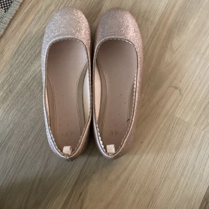 Glittriga ballerinaskor från H&M - Säljer ett par superglittriga ballerinaskor från H&M i storlek 37. De är i en snygg rosa nyans och perfekta för festliga tillfällen. Skorna har en rund tå och är i bra skick. Perfekta för att lägga till lite glitter i vardagen eller till en speciell kväll! ✨