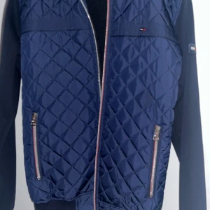 Blå quiltad jacka från Tommy Hilfiger - Snygg blå quiltad jacka från Tommy Hilfiger med dragkedja och långa ärmar. Perfekt för höst och vår. Den har en stilren design med små logodetaljer på bröstet och ärmen. Jackan är i mycket bra skick och passar perfekt för en casual look.
