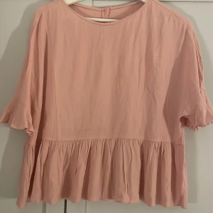 Rosa blus med volangdetaljer - Säljer en söt rosa blus med volangdetaljer i nederkant. Blusen har trekvartsärmar och knappar i ryggen för en fin detalj. Perfekt för vår och sommar! 🌸