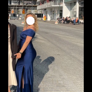 Marinblå långklänning med slits - Marinblå långklänning i ett glansigt material. Klänningen har offshoulder-design och en slits framtill. Använd 1 gång precis som ny.