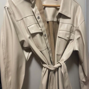 Beige trenchcoat med bälte - Säljer en snygg beige trenchcoat i syntetmaterial. Jackan har långa ärmar och knappar framtill. Den är utrustad med ett bälte i midjan för en elegant look. Perfekt för höst och vår!