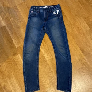 Blå jeans från Hampton Republic - Säljer ett par snygga blå jeans från Hampton Republic. De har en klassisk femficksdesign med coola detaljer som en liten amerikansk flagga på fickan och siffran 27 tryckt vid höften. Perfekta för vardagsbruk med en bekväm passform. Storlek 140cm. Använd ca 2 gånger.😊
