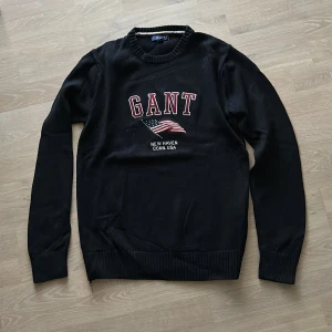 Svart stickad tröja från Gant strlk M - Säljer en snygg svart tröja från Gant med stickad logga på bröstet. Den har långa ärmar och en ribbad krage. Perfekt för en casual look eller när du vill ha något bekvämt men stilrent. Strlk M