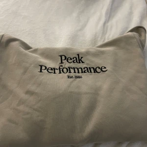 Beige hoodie från Peak Performance - Säljer en snygg beige hoodie från Peak Performance i storlek 170. Den är i bra skick och har en klassisk design med loggan tryckt på framsidan. Perfekt för både vardag och chill dagar. Huvan är mjuk och bekväm, och materialet känns skönt mot huden.