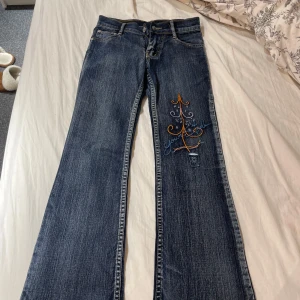 Broderade jeans med bootcut - Säljer ett par snygga bootcut jeans i blå denim med coolt broderi i orange och blått på ena benet. De har en klassisk femficksdesign och knappar i silverfärg. Perfekta för en avslappnad stil med en twist!