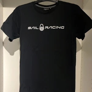 Svart t-shirt från Sail Racing - Snygg svart t-shirt från Sail Racing i bomull. Den har en klassisk passform med korta ärmar och ett stilrent vitt tryck på bröstet. Perfekt för en avslappnad stil eller för att visa upp ditt intresse för segling. Passar både till vardags och till mer sportiga aktiviteter.