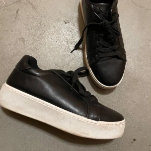 Svarta sneakers med vit sula - Säljer ett par svarta sneakers i skinn med vit sula. stilren design som passar perfekt till vardags. Skorna är i bra skick och redo för nya äventyr.