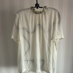 Vit blus  med dekorerad krage HM Ny - Säljer en vit blus med korta ärmar och en unik krage dekorerad med färgglada stenar och nitar i gult och turkost. HM, st.36 , Nytt!Perfekt för att ge en extra touch till din outfit. Passar både till vardags och festliga tillfällen.