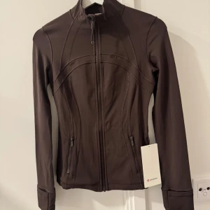 Brun jacka från Lululemon - Säljer en Nulu define jacket från Lululemon, helt ny med prislapp kvar. I färgen espresso, storlek 4. Kvitto finns 
