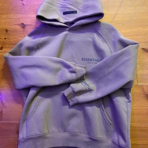 Beige hoodie från Essentials - Säljer en snygg beige hoodie från Essentials, Fear of God. Den är supermjuk och perfekt för kyliga dagar. Hoodien har en klassisk passform med ribbade muddar och en känguruficka framtill. Essentials-loggan är tryckt på bröstet och en liten patch finns på huvan. Perfekt för en avslappnad stil!