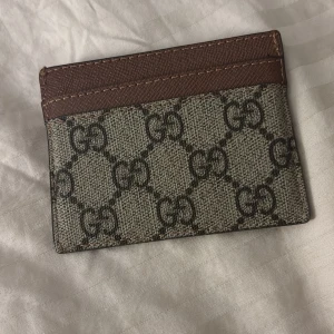 kortfodral med mönster - Säljer ett snyggt kortfodral i mycket bra skick. Det har ett klassiskt mönster i beige och brunt, med roliga färgglada figurer på ena sidan. Perfekt för att hålla dina kort organiserade med stil! Priset kan diskuteras 