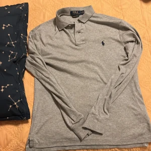 Grå pikétröja från Ralph Lauren - Säljer en stilren grå pikétröja från Ralph Lauren i custom slim fit. Tröjan är i bomull och har långa ärmar med en klassisk krage och knappar framtill. Perfekt för både vardag och lite finare tillfällen. Den är i mycket bra skick och redo för en ny ägare!