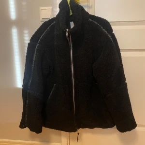 Svart teddyjacka från H&M som ny - En svart tjock och varm pilejacka i mjuk pile med kantband i ytbehandlad kvalitet. Jackan har lös passform, ståkrage, tvåvägsdragkedja fram och kängurufickor. Fodrad. Storlek: M (oversize) Skick: Använd 3 tillfällen och som ny.