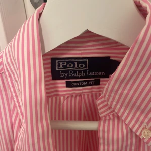 Rosa och vit randig skjorta från Ralph Lauren - Säljer en snygg rosa och vit randig skjorta från Ralph Lauren i custom fit. Skjortan har långa ärmar och är i mycket bra skick. Den är gjord av bomull och har en broderad blå logga på bröstet. Perfekt för både vardag och fest! 💖