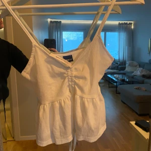 Vitt linne med snörning - Säljer en söt vit linnetopp. Den har smala axelband och en knytning framtill som ger en fin detalj. Perfekt för sommardagar eller att styla med en kofta för kyligare kvällar. Toppen är i ett lätt och luftigt material.