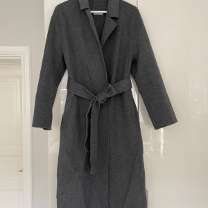 Grå ullkappa med bälte - Säljer en elegant grå kappa i ull med knytbälte i midjan. Perfekt för höst och vinter, med långärmad design och klassisk krage. Kappan är i mycket bra skick och ger en stilren look. Passar både till vardags och festliga tillfällen.