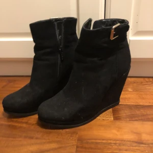 Svarta kilklackade boots med dragkedja - Snygga svarta boots med kilklack från Jenny Fairy. De har en praktisk dragkedja på sidan och en dekorativ spänne upptill. Perfekta för höst och vinter, dessa boots är både stiliga och bekväma. Passar bra till både jeans och klänning! 