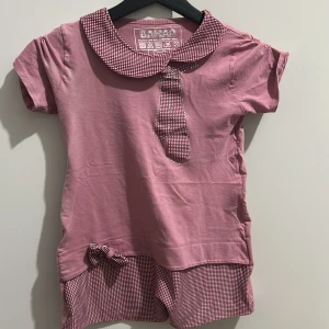 Rosa topp från Bahar - Söt rosa topp från Bahar med korta ärmar och ett rutigt mönster på kragen och nederkanten. Den har en dekorativ rosett och en knytning framtill. Perfekt för en avslappnad dag eller en somrig utflykt!