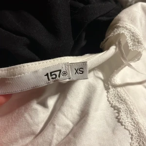 Vit topp med spetsdetaljer från 157 - Säljer en söt vit topp från 157 i storlek XS. Den har korta ärmar och fina spetsdetaljer vid halsringningen. Toppen är tight och har en liten rosett framtill. Perfekt för sommardagar eller en avslappnad kväll med vänner.
