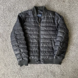 Svart pufferjacka från Polo Ralph Lauren - Säljer en snygg svart pufferjacka från Polo Ralph Lauren. Jackan är perfekt för kyliga dagar med sin quiltade design och dragkedja framtill. Den har en klassisk look med ribbade muddar vid ärmslut och nederkant. Passar både till vardags och lite finare tillfällen. 🖤