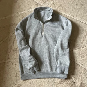 Grå fleece tröja med dragkedja - Mysig grå fleece tröja perfekt för kyliga dagar. Den har en halv dragkedja framtill och långa ärmar med ribbade muddar. Enkel och stilren design som passar till det mesta. Perfekt för både vardag och avslappnade tillfällen.