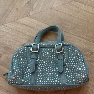 Grå axelväska med strassdetaljer - Snygg grå axelväska i jeansmaterial med glittrande strassdetaljer över hela väskan. Den har två handtag och en justerbar axelrem. Perfekt för att ge lite extra bling till din outfit! Väskan stängs med dragkedja och har en stilren design som passar både till vardags och fest.