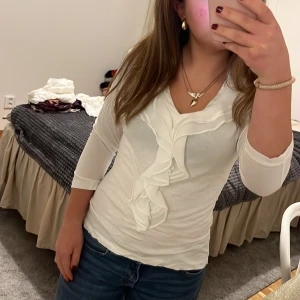 Vit blus med volangdetaljer - Säljer en stilren vit blus med v-ringning och volangdetaljer framtill. Blusen har trekvartsärmar och är perfekt för både vardag och fest. Den är i mycket bra skick och passar till många olika outfits.