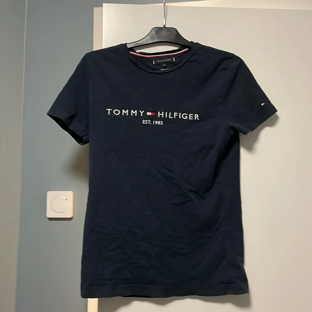 Säljer en snygg mörkblå t-shirt från Tommy Hilfiger i ekologisk bomull. Den har korta ärmar och ett stilrent tryck med märkesnamnet och 'Est. 1985' på framsidan. Perfekt för en casual look!. T-shirts.