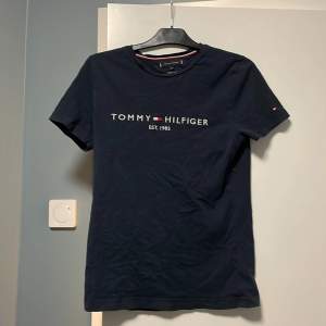 Säljer en snygg mörkblå t-shirt från Tommy Hilfiger i ekologisk bomull. Den har korta ärmar och ett stilrent tryck med märkesnamnet och 'Est. 1985' på framsidan. Perfekt för en casual look!