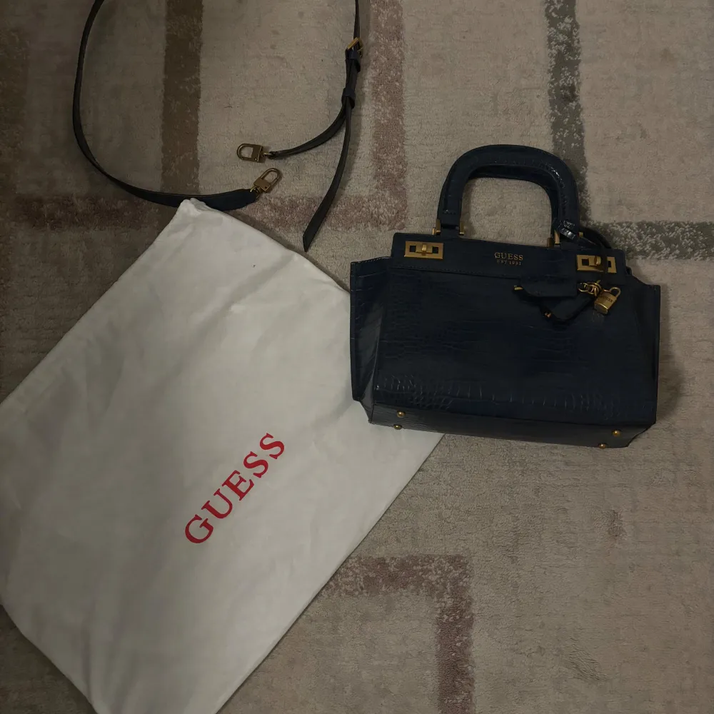 Säljer en elegant navy handväska från Guess som är helt ny. Väskan har en krokodilpräglad yta och guldfärgade detaljer. Den har både handtag och en avtagbar axelrem. Insidan är fodrad med Guess-logotyp och har flera fack med dragkedja. Perfekt för både vardag och fest!. Väskor.