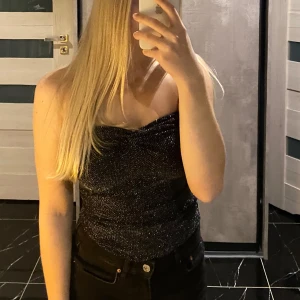 Glittrig tubtopp - Snygg glittrig tubtopp perfekt för fest! Den är svart med silverglitter och har en åtsittande passform. Toppen är axelbandslös och har en elegant twistdetalj framtill. Perfekt för att glamma upp vilken outfit som helst!