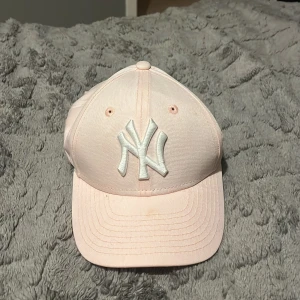 Ljusrosa keps från New Era - Säljer en ljusrosa keps från New Era med broderad logga framtill. Kepsen är i bomull och har en justerbar rem baktill. Den är i bra skick men har en liten fläck på skärmen. Perfekt för sommarens äventyr! 🌸