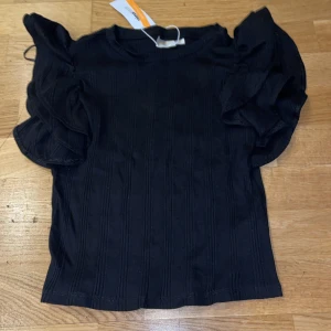 Svart topp med volangdetaljer - Snygg svart topp med volangdetaljer på ärmarna. Perfekt för både vardag och festliga tillfällen. Toppen har en ribbad struktur som ger en extra touch av stil. Ny med taggar kvar!