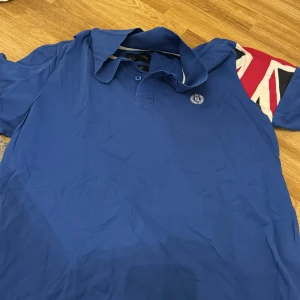 Blå pikétröja från Henri Lloyd - Säljer en snygg blå pikétröja från Henri Lloyd i bra skick. Tröjan har en klassisk krage och knappar framtill. Den har ett stilrent märke på bröstet och är perfekt för både vardag och fest. Passar bra till jeans eller chinos!