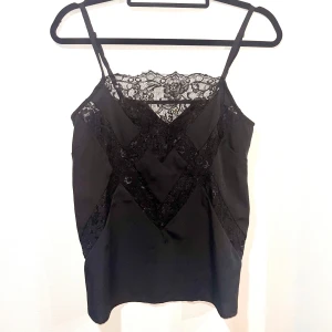 Svart linne med spetsdetaljer - Snygg svart linne med tunna axelband och vacker spetsdetalj över bysten och magen. Perfekt för en utekväll eller fest. Materialet känns mjukt och lyxigt. 