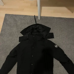 Canada goose black label - Säljer en oanvänd svart canada goose dunjacka  med huva och knappar. Jackan har en broderad logga på ärmen och är perfekt för kalla dagar. Den kommer med en praktisk förvaringsväska.