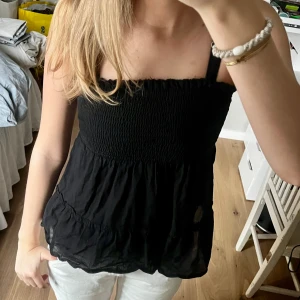 Svart axelbandslös topp - Snygg svart axelbandslös topp med smockdetaljer upptill och volang nedtill. Perfekt för varma dagar och kan enkelt matchas med olika outfits.