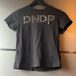 Svart t-shirt från Dondup - Snygg svart t-shirt från Dondup med glittrande detaljer på framsidan som bildar bokstäverna DNDP. Tillverkad i Italien, perfekt för en stilren look. Tröjan saknar en del av dom vita tofsarna i loggan, sen finns det ett litet tvättmasinshål på baksidan bredvid D-et, står ingen storlek på den men skulle säga att det är en xss, jag hade den när jag var ca 10 år och jag var ett litet barn eller hur jag skall säga det. Frakt betalar köparen, kan hämtas i Mölnlycke.