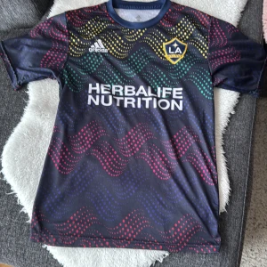 LA Galaxy fotbollströja med färgglatt mönster - Snygg LA Galaxy fotbollströja från Adidas med ett vågigt, färgglatt mönster i rött, gult och grönt. Tröjan har korta ärmar och klubbens emblem på bröstet. Perfekt för fans av laget! Storlek 164, barnstorlek 