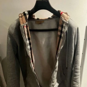 Grå hoodie från Burberry - Säljer en stilren grå hoodie från Burberry med klassiskt rutigt mönster i huvan. Tröjan har dragkedja framtill och broderad logga på bröstet. Perfekt för en avslappnad och trendig look. Storlek M men funkar även för S. Använd Max 5 gånger