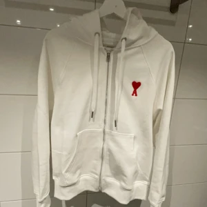 Vit hoodie från Ami - Säljer en stilren vit hoodie från Ami med en röd hjärtlogga på bröstet. Den har dragkedja framtill och justerbar huva med snören. Perfekt för en avslappnad look.