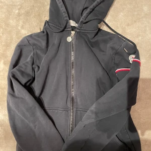 Svart huvtröja från Moncler - Säljer en stilren svart huvtröja från Moncler med dragkedja och justerbar huva. Tröjan har en broderad logotyp på ärmen och ribbade muddar. Perfekt för en casual look. Tror den är äkta, skriv för fler bilder. Vet alltså ej så ingen garanti på att den är de